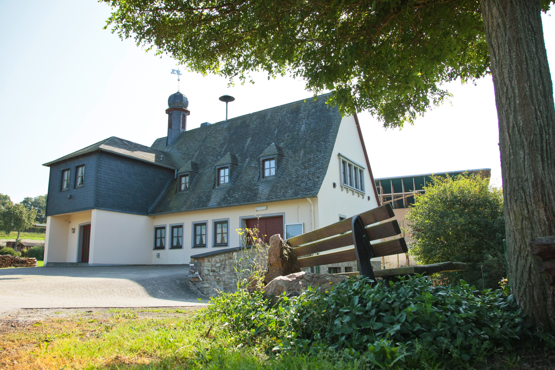 Gemeindehaus der OG Bergenhausen, im Vordergrund ist eine Holzbank unter Bäumen