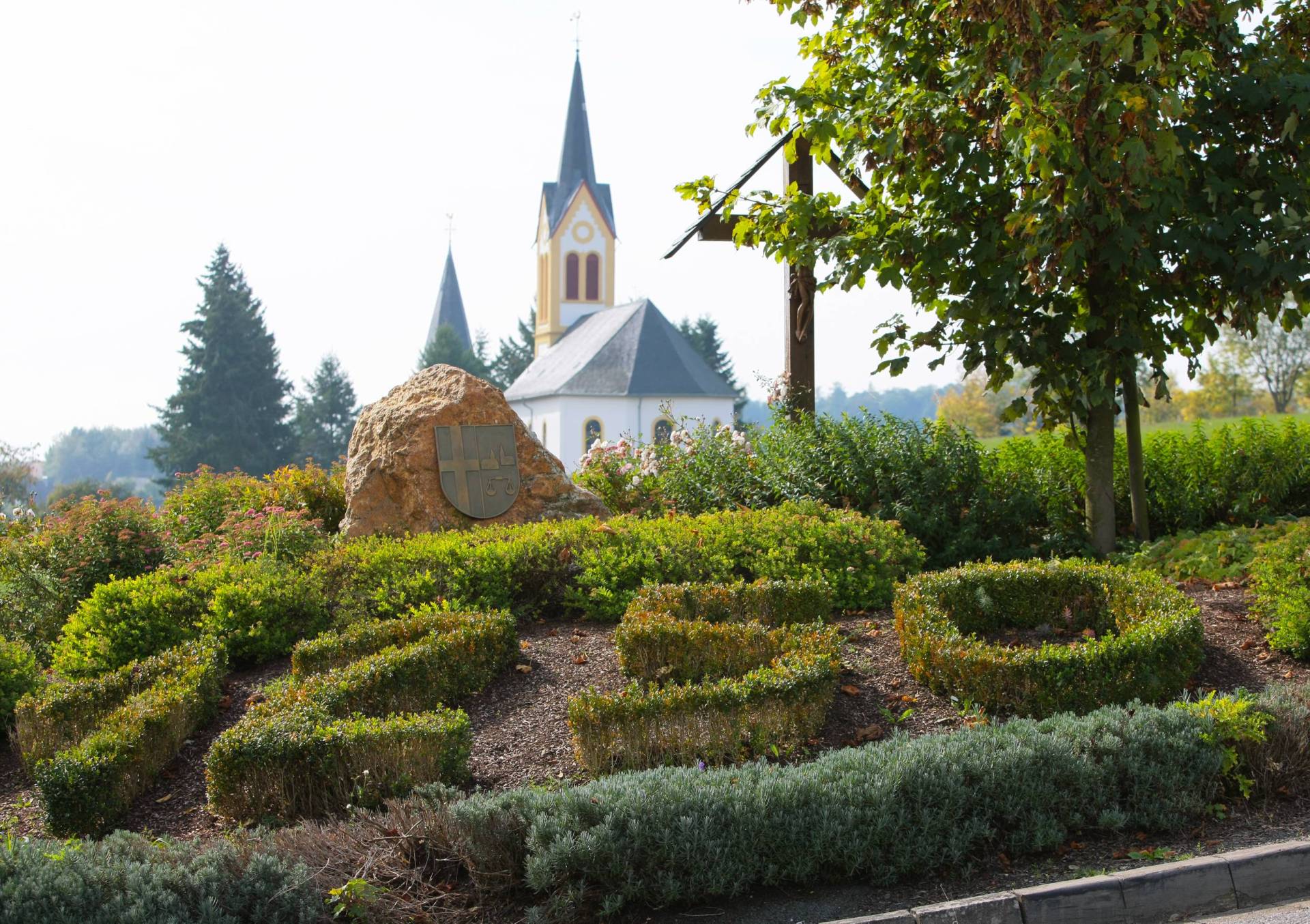 Blick auf die Kirche in Biebern, im Vordergrund ist ein Beet mit Sträuchern und auf einem im Beet liegenden Stein ist das Wappen der Ortsgemeinde aufgesetzt
