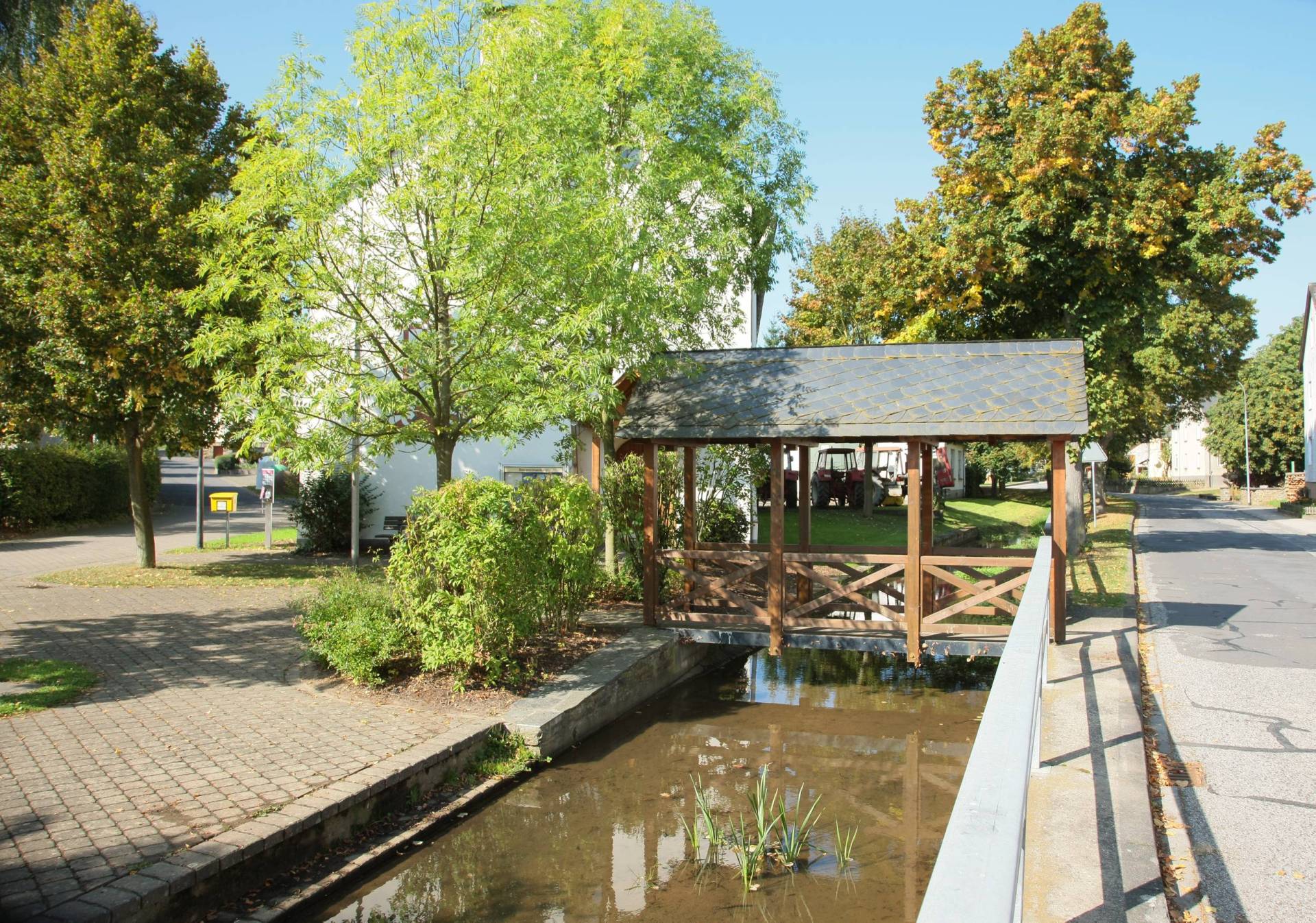 Dorfmitte mit einer Brücke über den Bach inmitten von Bäumen bei blauem Himmel und strahlendem Sonnenschein