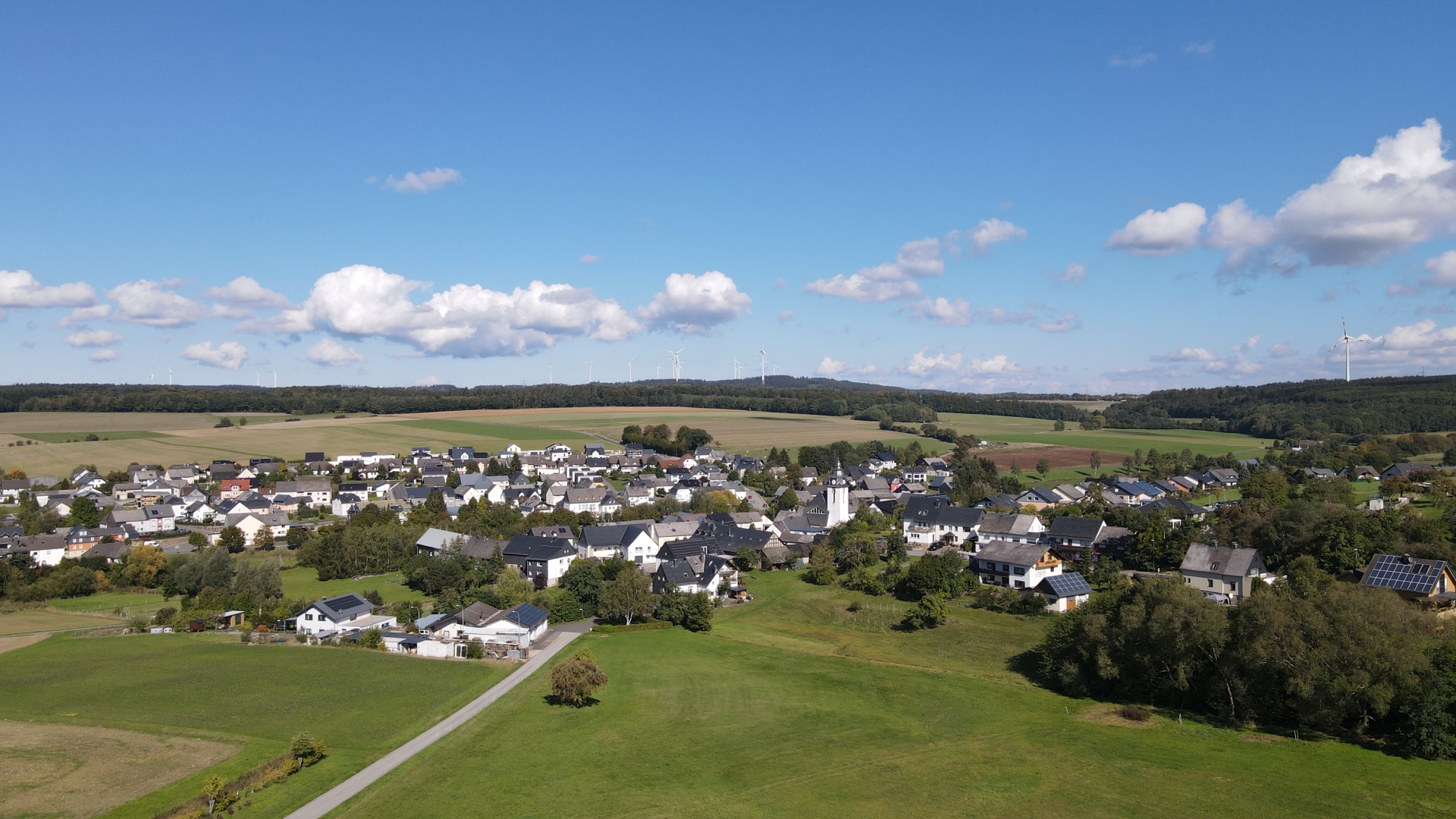 Drohnenaufnahme Ortsansicht von Kisselbach mit Windrädern im Hintergrund bei blauem Himmel