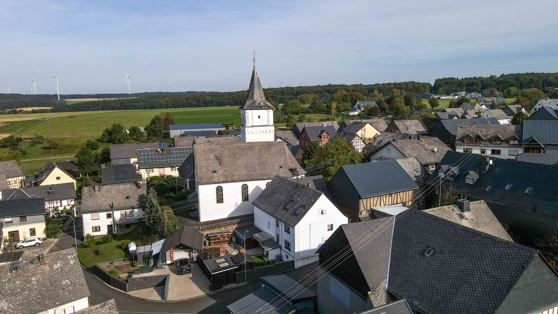 Häuseransicht mit der Kirche im Dorfmittelpunkt, im Hintergrund der Wald mit Windrädern