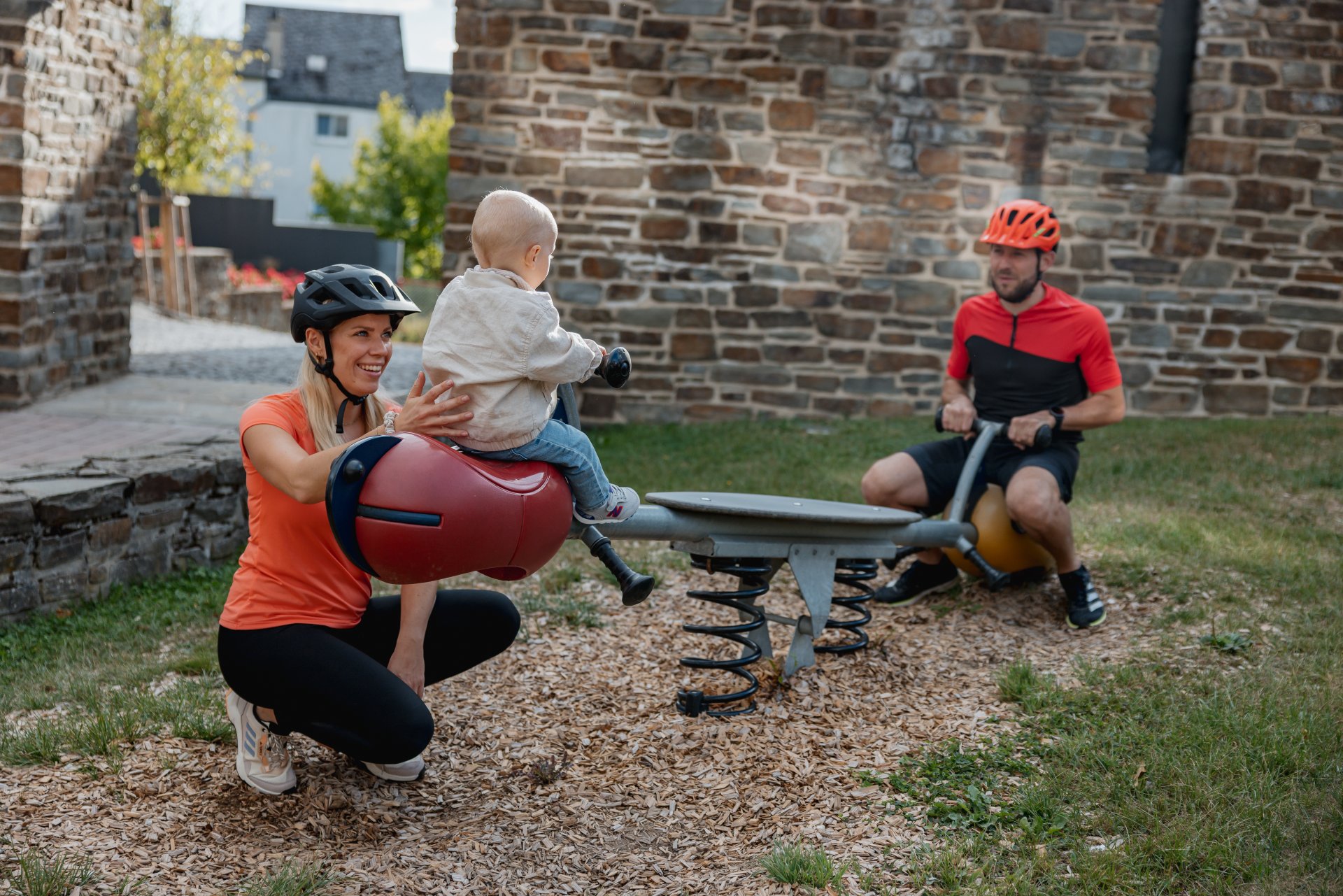 Ein kleines Kind sitzt auf einer Wippe mit dem Vater und die Mutter hält das Kind fest.