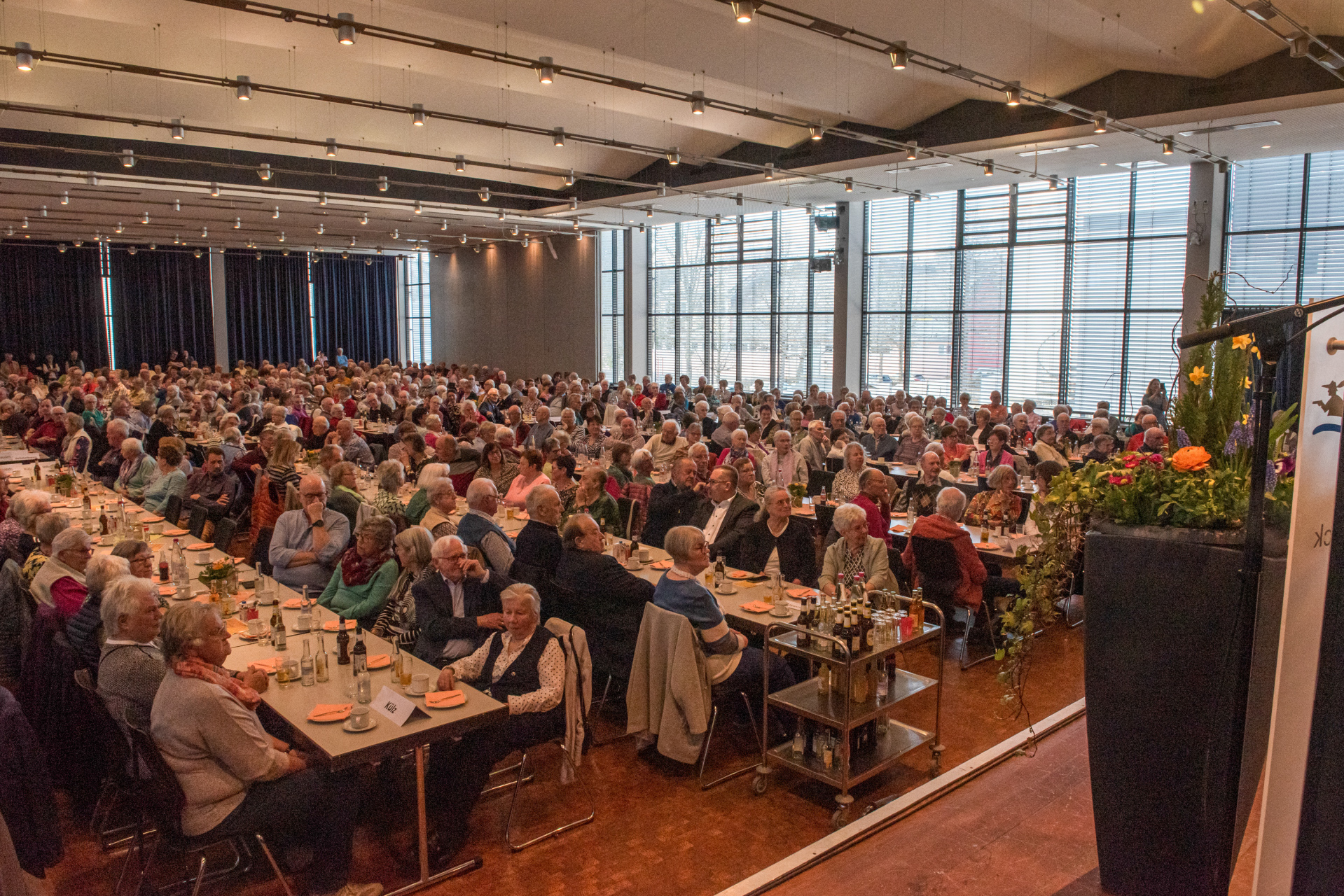 Auf dem Bild sitzen 600 Seniorinnen und Senioren in der Hunsrückhalle Simmern an Tischen mit dem Blick Richtung Bühne. Die Tische sind in Reihenform gestellt und eingedeckt. 
