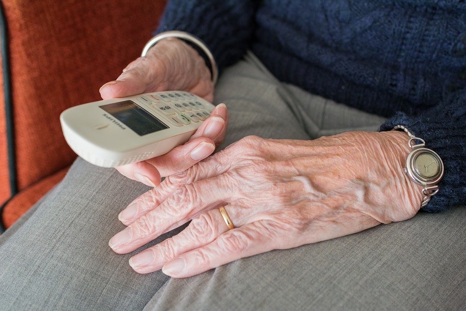 Auf dem Bild sind die Hände einer älteren Person zu sehen. Sie hat die Hände in ihren Schoß gelegt und hält in der einen Hand ein schnurloses, helles Telefon.