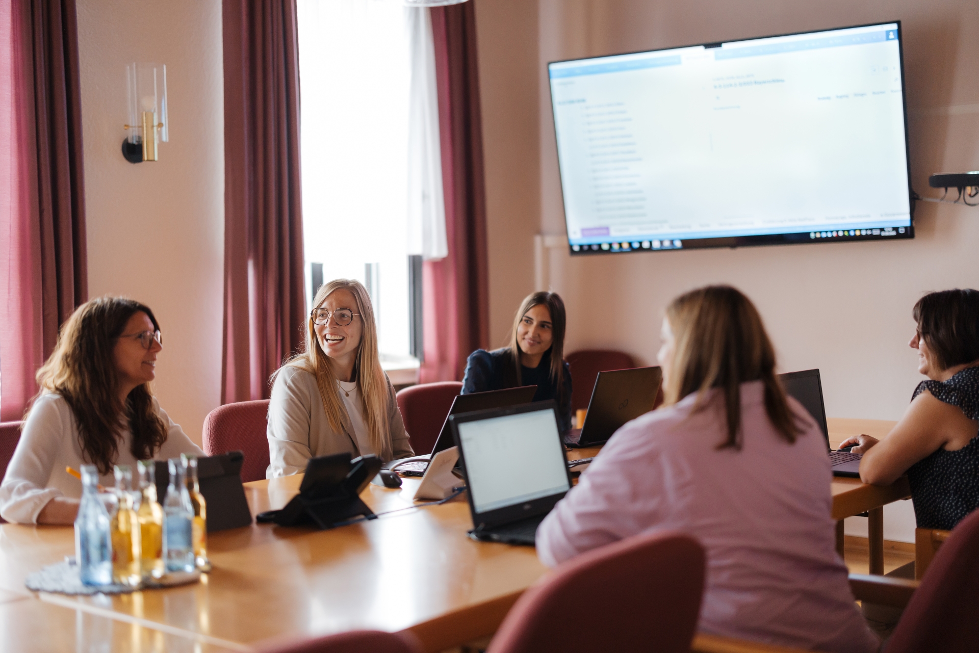 Mitarbeiterinnen der Verbandsgemeindeverwaltung sitzen im Sitzungssaal und halten eine Besprechung ab. Vor ihnen steht jeweils ein Laptop. 