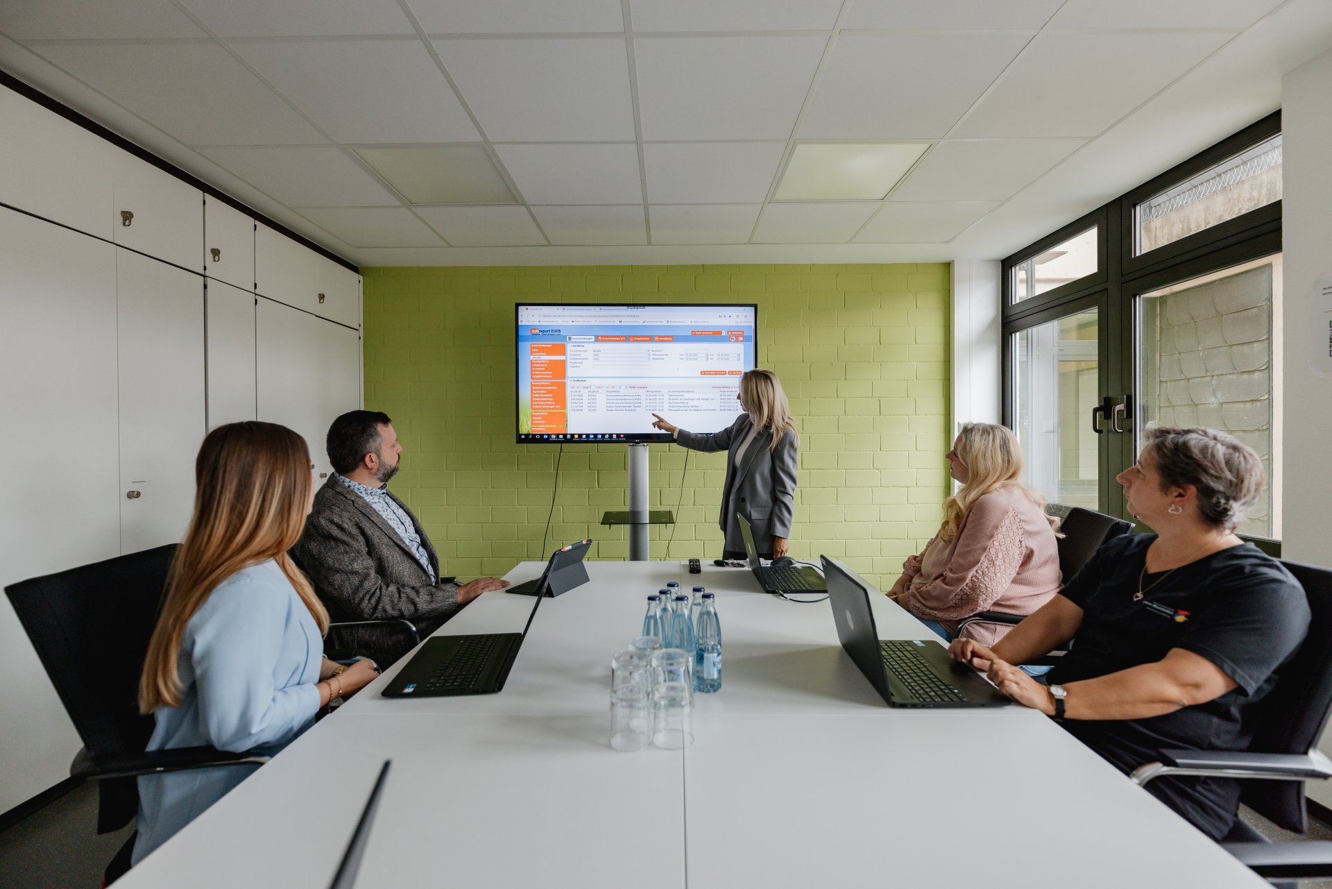 Mehrere Personen sitzen im Sitzungssaal zusammen. Eine weitere Person präsentiert die öffentliche Azsschreibung auf einem großen BIldschirm