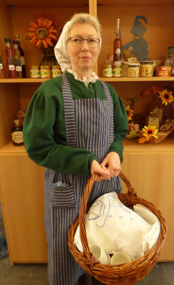 Eine Frau steht verkleidet mit enem Kopftuch und einer Schürze und einem Korb mit Tassen vor einem Regal mit Hunsrücker Regionalitäten.