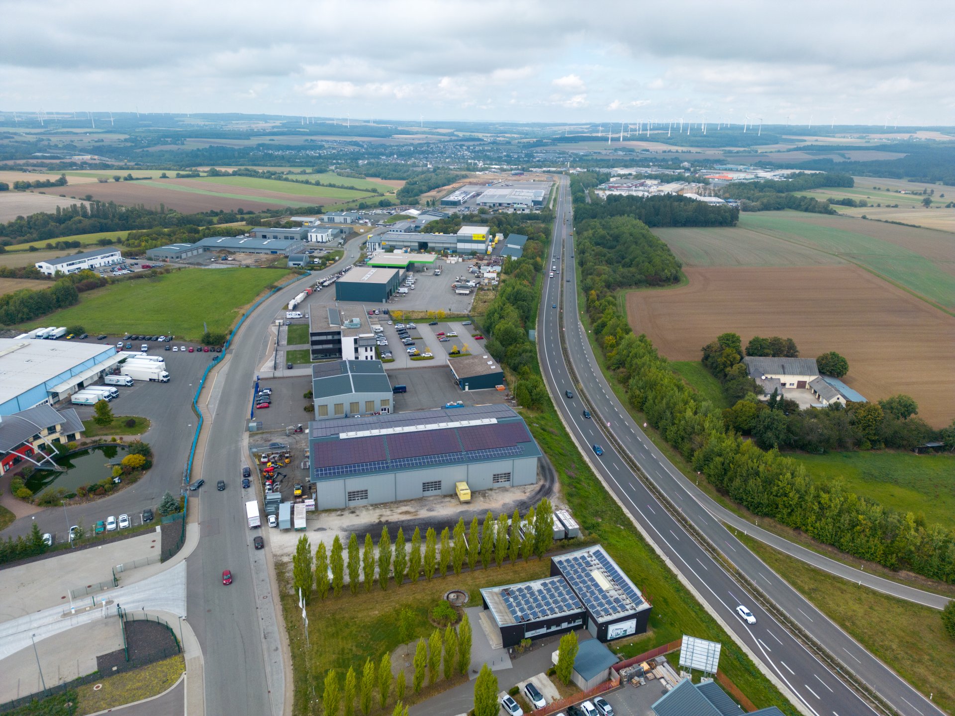 Luftaufnahme des Industrieparks Simmern im Hunsrück mit Gewerbehallen, Parkflächen, der vorbeiführenden B50 sowie umliegenden Feldern und Waldflächen im Hintergrund.