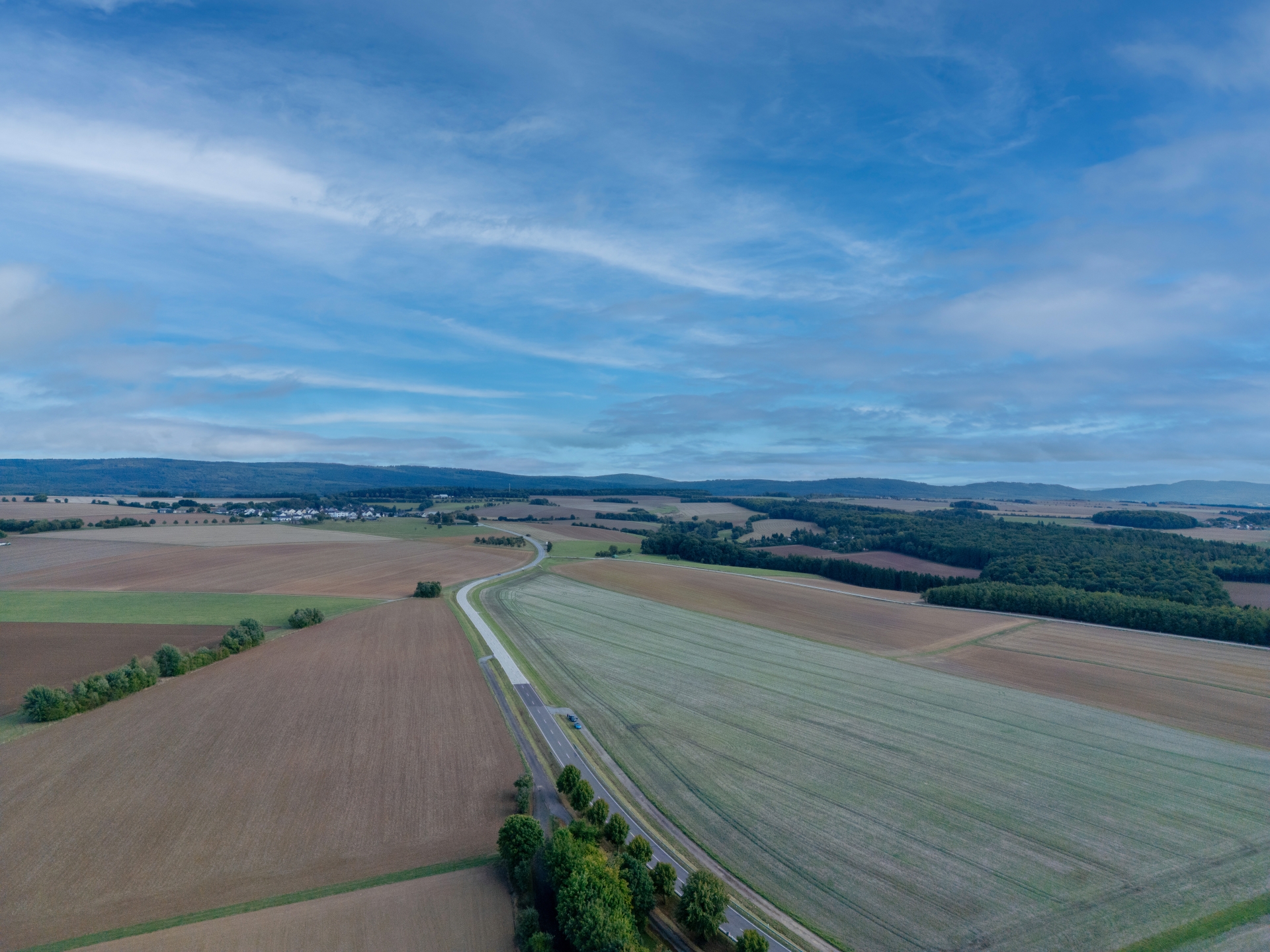 Luftaufnahme landwirtschaftlicher Flächen in der Verbandsgemeinde Simmern-Rheinböllen im Hunsrück mit Feldern, Baumstreifen und einer Landstraße unter bewölktem Himmel.