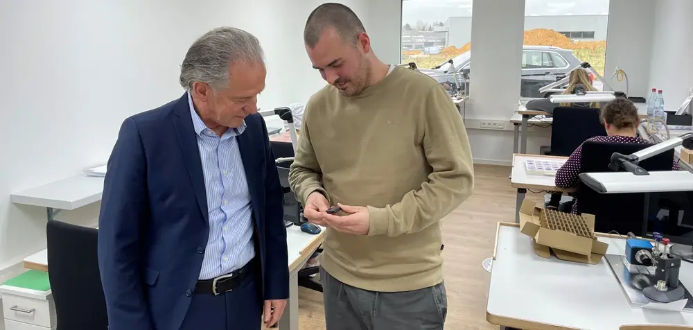 Zwei Personen stehen in einem Büro und betrachten gemeinsam einen kleinen Gegenstand, im Hintergrund sind Arbeitsplätze und weitere Personen an Schreibtischen sichtbar.