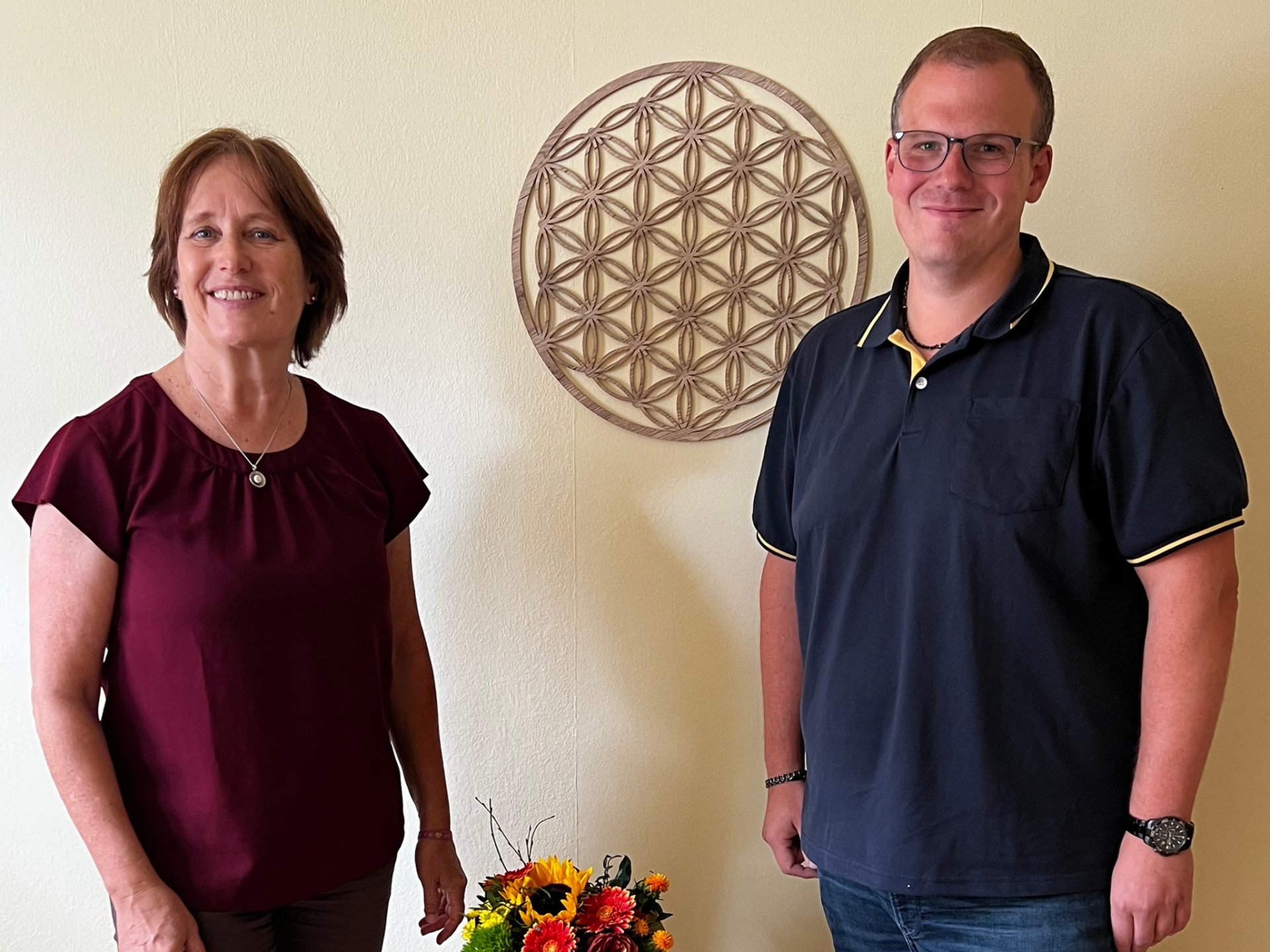 Foto - Unternehmensbesuch Balance Gottschalk Zwei Personen (Uta Gottschalk von Balance Gottschalk und Kai Quint von der Wirtschaftsförderung der Verbandsgemeinde Simmern-Rheinböllen) stehen lächelnd vor einer Wand mit floralem Dekor. Vor ihnen ein kleiner Strauß mit bunten Blumen. In den Praxisräumen von Balance Gottschalk in Rheinböllen.
