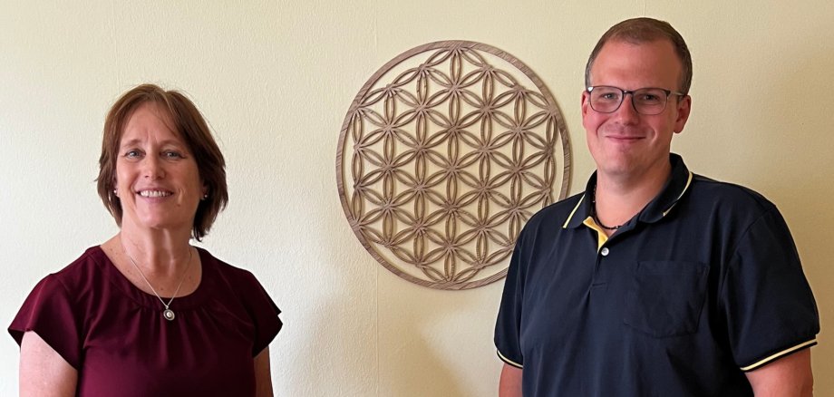 Foto - Unternehmensbesuch Balance Gottschalk Zwei Personen (Uta Gottschalk von Balance Gottschalk und Kai Quint von der Wirtschaftsförderung der Verbandsgemeinde Simmern-Rheinböllen) stehen lächelnd vor einer Wand mit floralem Dekor. Vor ihnen ein kleiner Strauß mit bunten Blumen. In den Praxisräumen von Balance Gottschalk in Rheinböllen.