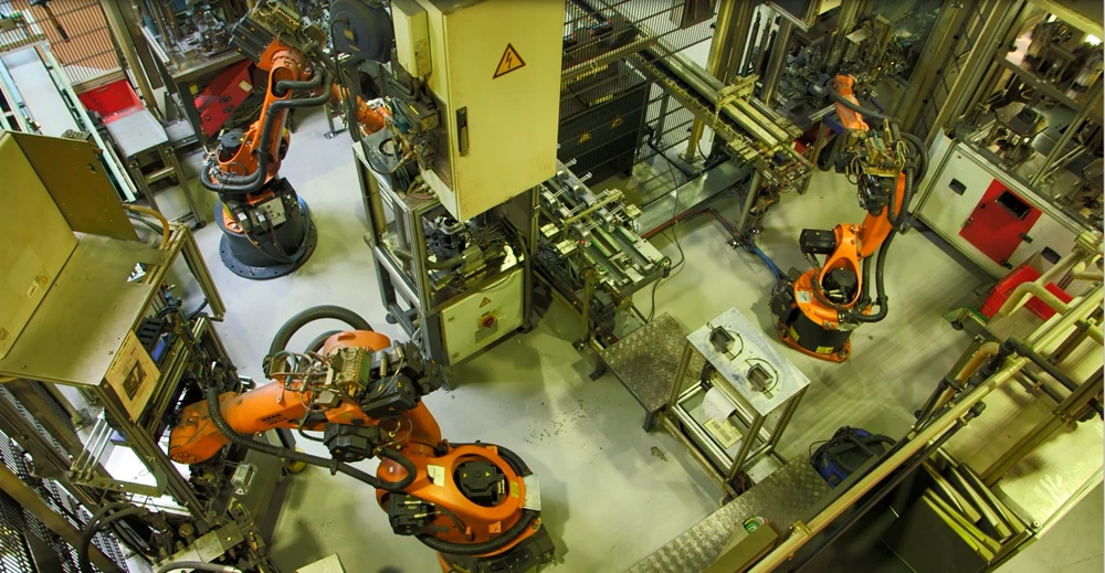 Industrielle Fertigungsanlage mit mehreren orangefarbenen Robotergreifarmen und automatisierten Produktionsstationen in einer Werkhalle.