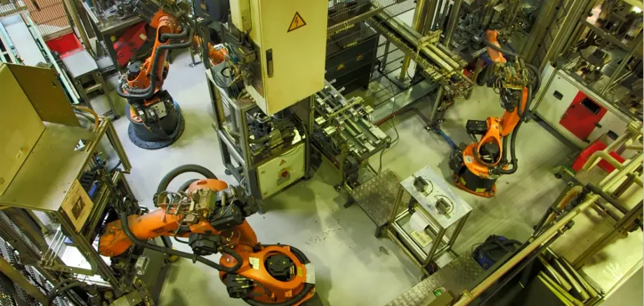 Industrielle Fertigungsanlage mit mehreren orangefarbenen Robotergreifarmen und automatisierten Produktionsstationen in einer Werkhalle.