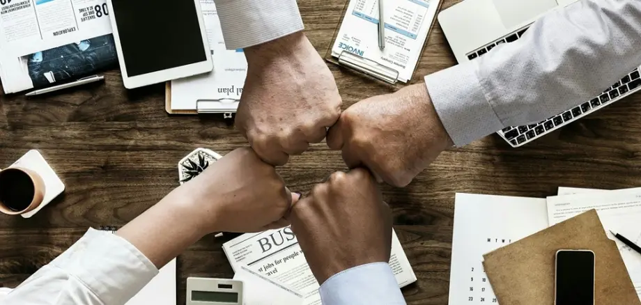 Vier Hände unterschiedlicher Personen stoßen in einer Bürosituation mit einem „Fist Bump“ über einem Tisch zusammen. Auf dem Tisch liegen Arbeitsunterlagen, Notizblöcke, ein Taschenrechner, ein Laptop und Dokumente. Symbolbild für Teamwork und Zusammenarbeit.