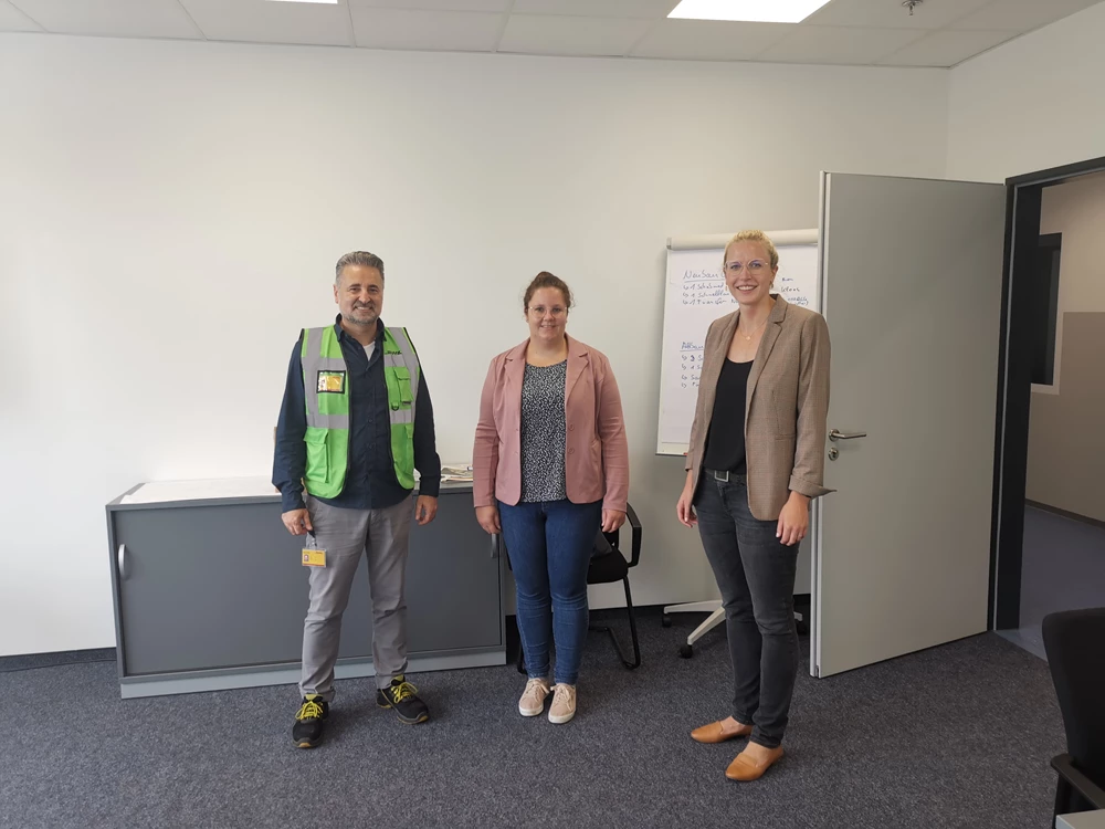 Unternehmensbesuch bei DHL Solutions am Standort Simmern im Rhein-Hunsrück-Kreis. Im Bild stehen drei Personen in einem Besprechungsraum nebeneinander vor einer weißen Wand und einem Flipchart: eine Person mit DHL-Sicherheitsweste, daneben zwei Besucherinnen. Der Raum wirkt hell, modern und funktional eingerichtet.