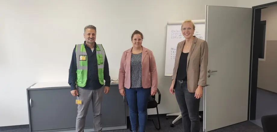 Unternehmensbesuch bei DHL Solutions am Standort Simmern im Rhein-Hunsrück-Kreis. Im Bild stehen drei Personen in einem Besprechungsraum nebeneinander vor einer weißen Wand und einem Flipchart: eine Person mit DHL-Sicherheitsweste, daneben zwei Besucherinnen. Der Raum wirkt hell, modern und funktional eingerichtet.
