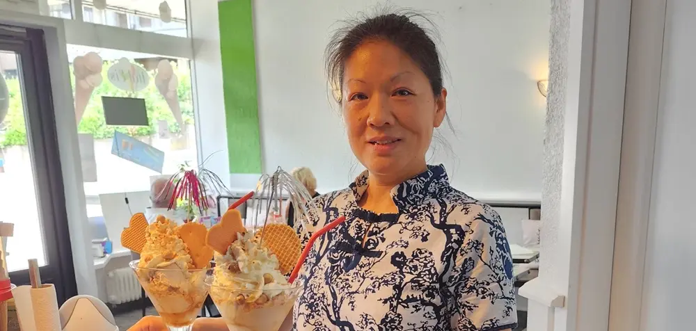 Person in einem Eiscafé hält ein Tablett mit zwei Eisbechern, die mit Sahne, Waffeln und Dekoration serviert sind, im Innenraum mit Sitzbereich im Hintergrund.