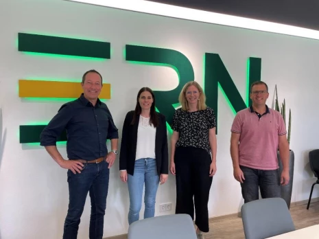 Vier Personen stehen in einem Büro vor einer Wand mit großem Schriftzug „ERN“, Sitzmöbel und Pflanzen im Hintergrund.
