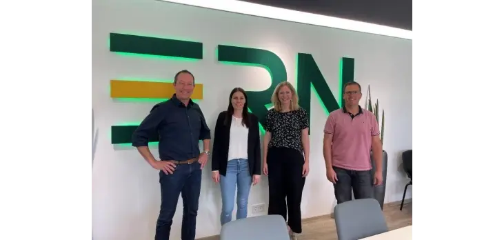 Vier Personen stehen in einem Büro vor einer Wand mit großem Schriftzug „ERN“, Sitzmöbel und Pflanzen im Hintergrund.