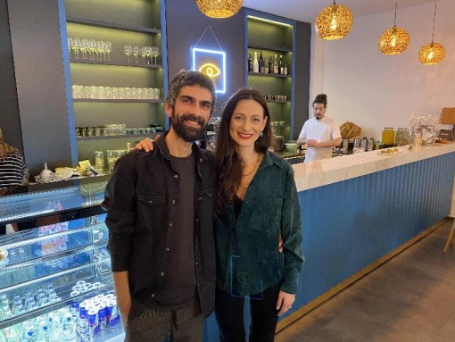 Zwei Personen stehen im Innenraum eines Cafés vor der Theke, daneben sind Glasregale, Getränke und ein weiterer Mitarbeitender im Hintergrund zu sehen.