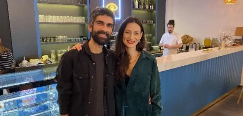 Zwei Personen stehen im Innenraum eines Cafés vor der Theke, daneben sind Glasregale, Getränke und ein weiterer Mitarbeitender im Hintergrund zu sehen.