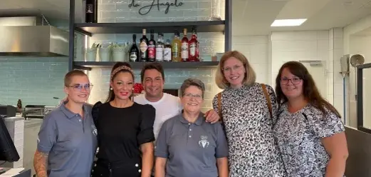 Personengruppe steht im Eingangs- und Tresenbereich des Restaurants „GiGi’s“, hinter ihnen sind Regale mit Flaschen, Gläsern und Küchenbereich sichtbar.