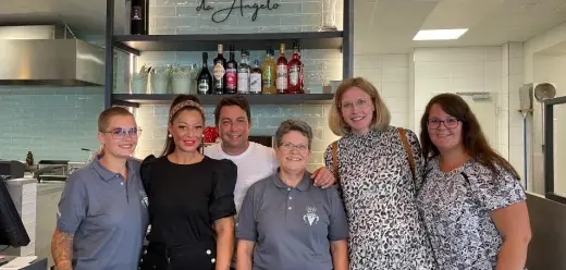 Personengruppe steht im Eingangs- und Tresenbereich des Restaurants „GiGi’s“, hinter ihnen sind Regale mit Flaschen, Gläsern und Küchenbereich sichtbar.