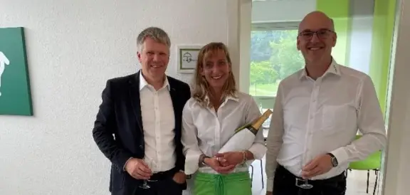 Drei Personen stehen in einem Innenraum nebeneinander und halten jeweils Gläser, im Hintergrund ist ein Raum mit Sitzmöbeln und Fenstern sichtbar.