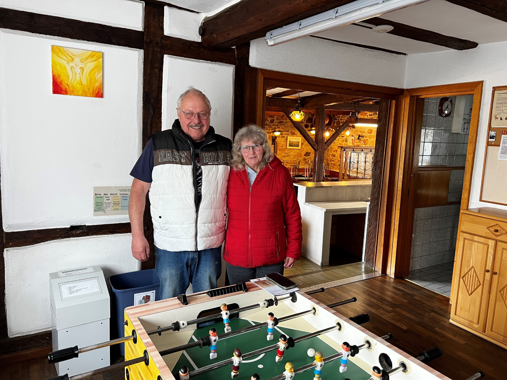 Zwei Personen (Dörthe Kauth, Siegfried Kauth) stehen lächelnd in einem gemütlichen Raum neben einem Tischfußballtisch, Holzbalken sichtbar. Im Freizeithaus Engel im Stadtteil Kleinweidelbach von der Stadt Rheinböllen im Hunsrück.