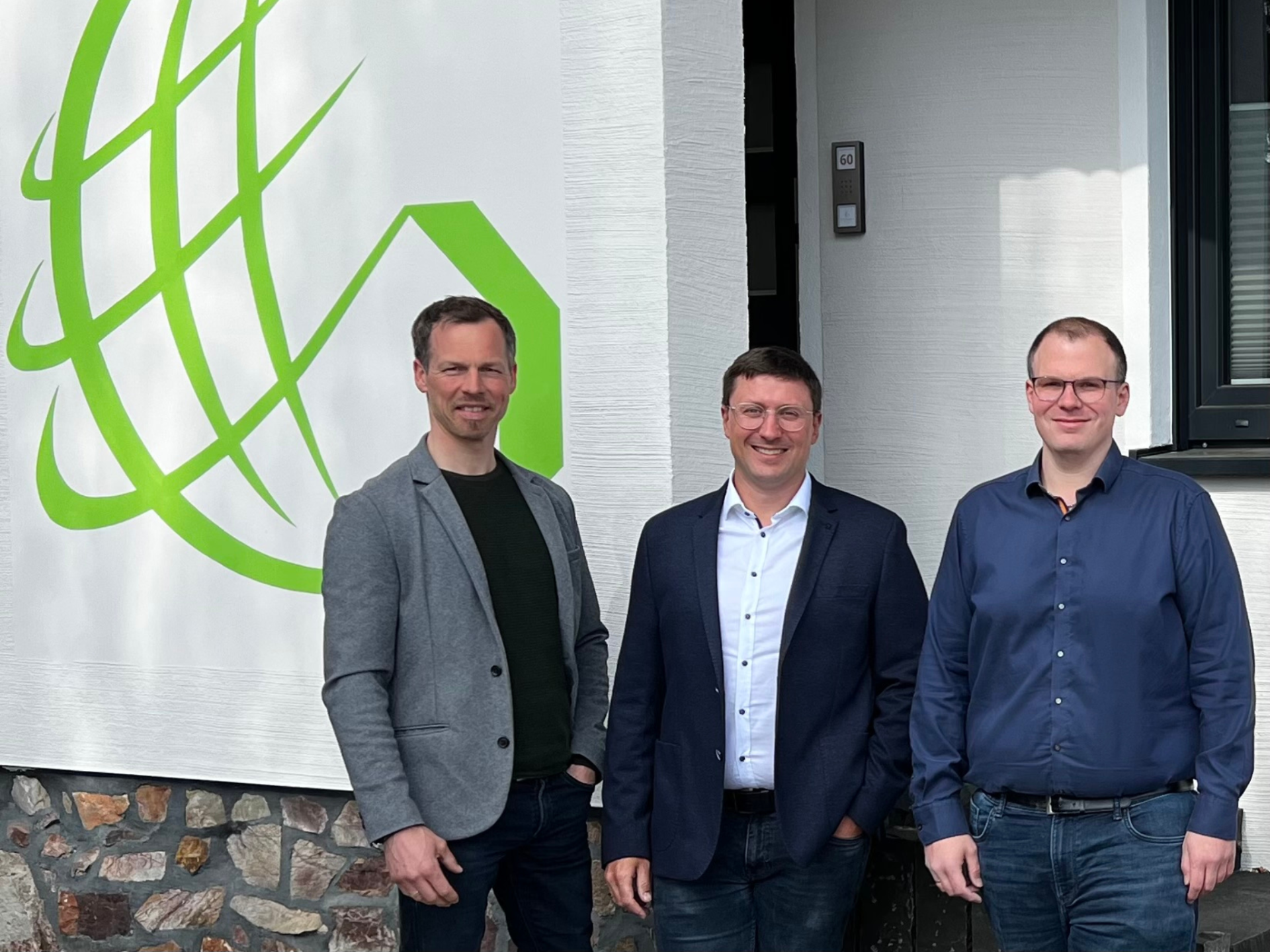 Drei Männer stehen lächelnd vor einem Gebäude, neben dem grünen Logo der GRÜNwerk GmbH an der Wand des Unternehmensgebäudes in Simmern/Hunsrück.