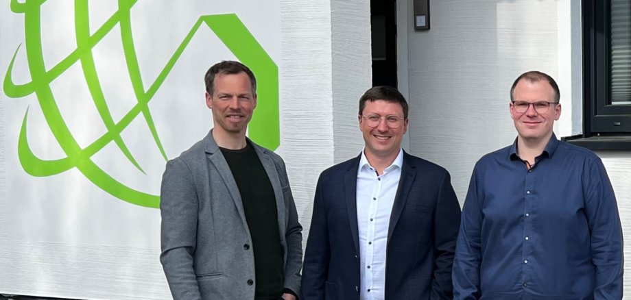 Fotos - GRÜNwerk GmbH Drei Männer stehen lächelnd vor einem Gebäude, neben dem grünen Logo der GRÜNwerk GmbH an der Wand des Unternehmensgebäudes in Simmern/Hunsrück.