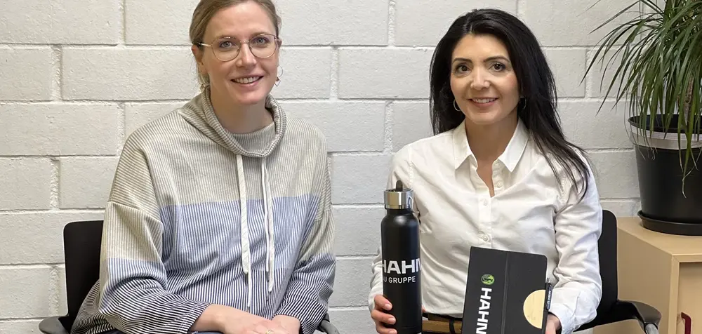 Zwei Personen sitzen nebeneinander vor einer weißen Innenwand; eine Person von Koby. hält eine Trinkflasche und eine Verpackung mit dem Logo „HAHN Gruppe“.