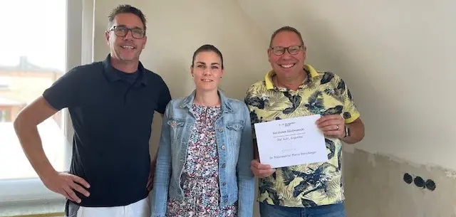 Drei Personen stehen in einem renovierungsbedürftigen Raum nebeneinander; eine Person hält ein Papier in den Händen, im Hintergrund sind Putzarbeiten und frei liegende Wandflächen erkennbar.