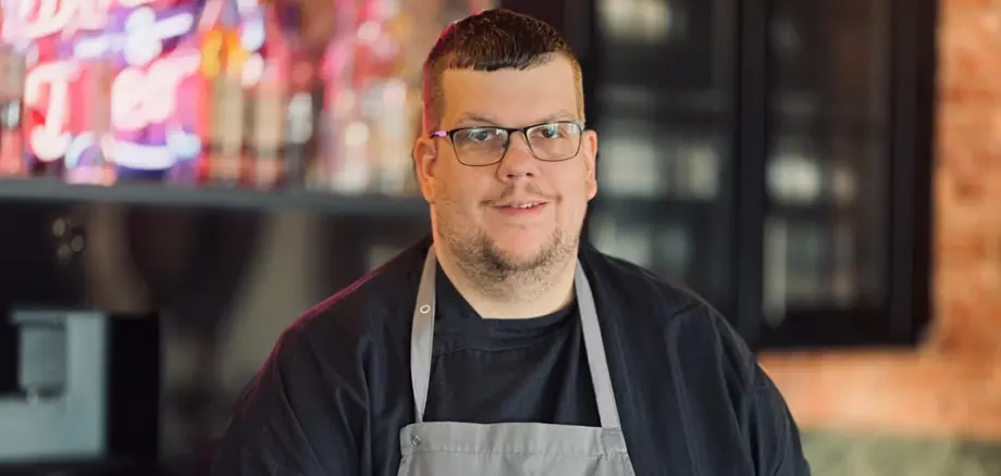 Person in Kochkleidung steht in einer Küche und trägt eine Schürze mit grafischem Aufdruck, im Hintergrund sind Kücheninventar und Beleuchtungselemente zu sehen.