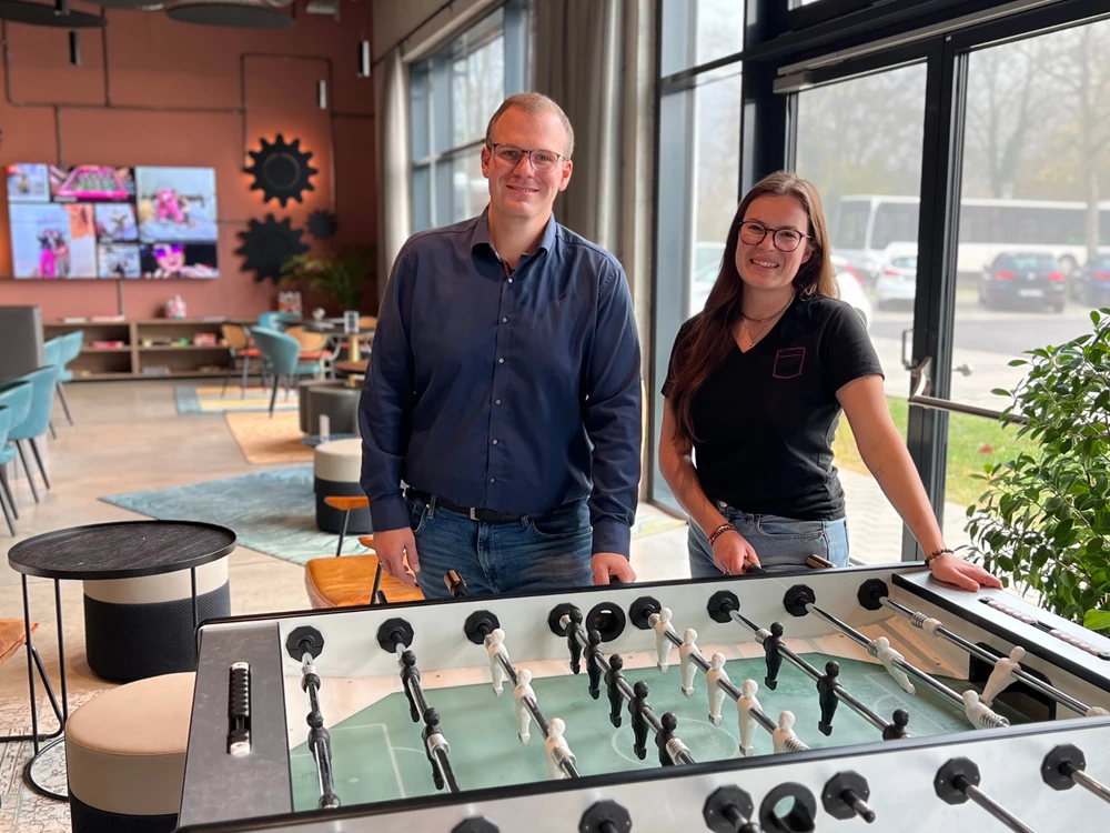 Zwei Personen stehen neben einem Tischkicker im Aufenthalts- und Empfangsbereich des Hotels Moxy in Simmern mit großen Fenstern und Sitzmöbeln im Hintergrund.