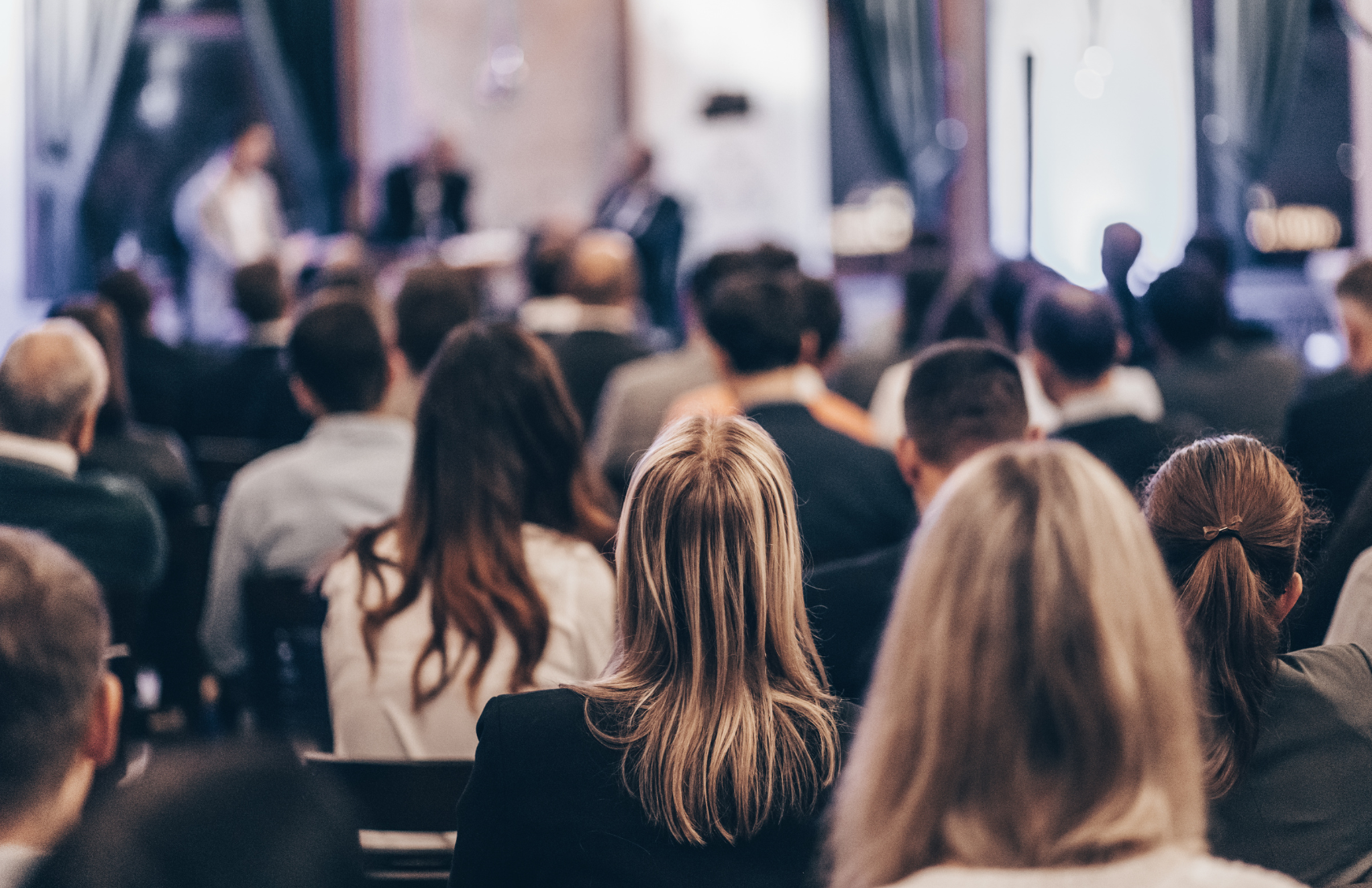 Publikum in einem Veranstaltungsraum, sitzend und nach vorn zur Bühne ausgerichtet, mit unscharf dargestellter Vortragssituation im Hintergrund.